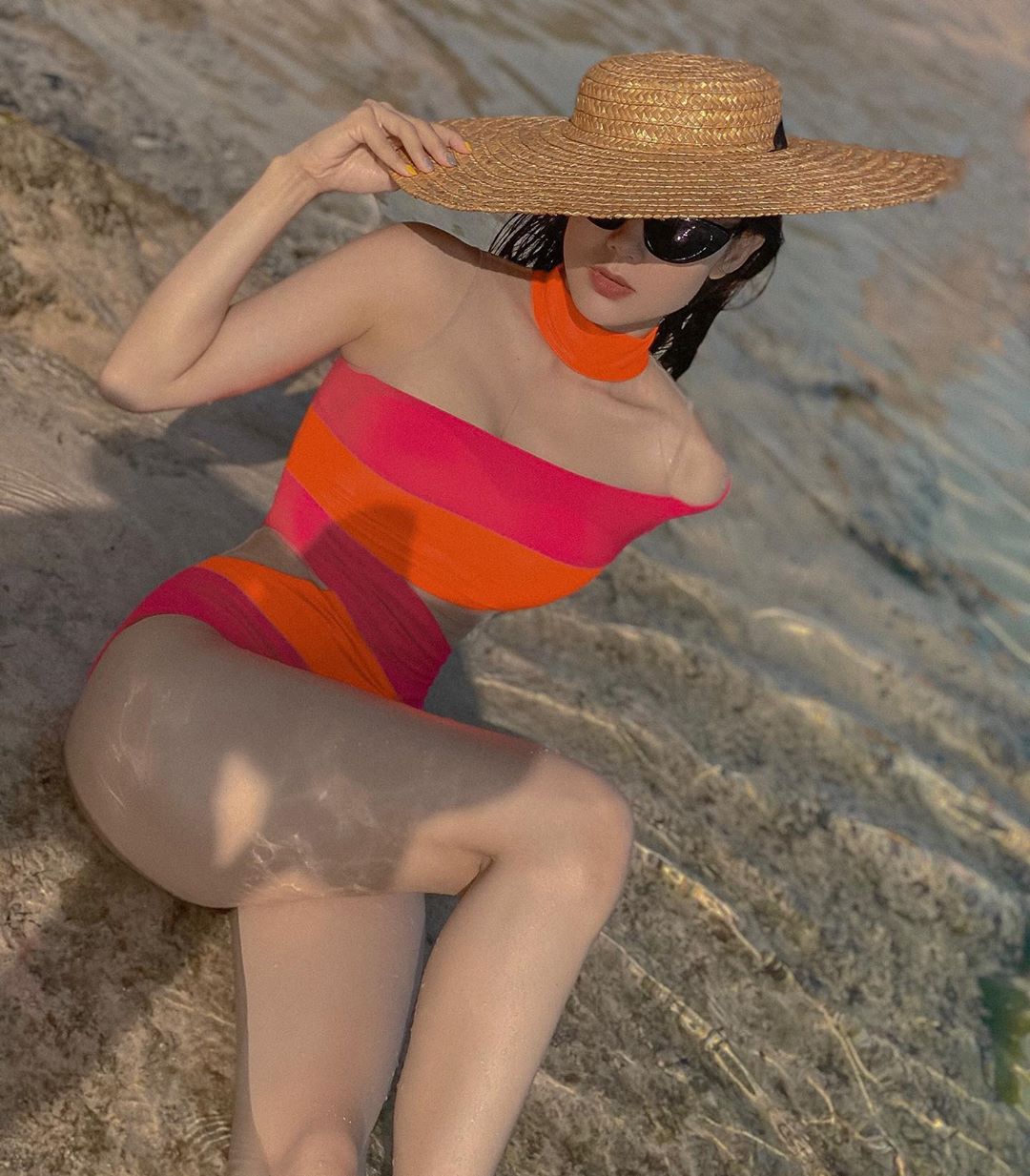 Minh Hằng mặc thiết kế monokini cut - out ở phần eo phối hai màu hồng và cam theo kiểu váy băng cuốn. Kết hợp với trang phục, Minh Hằng chọn kính mắt to bản màu đen cùng mũ cói rộng vành mang đậm không khí mùa hè và phù hợp đi biển.