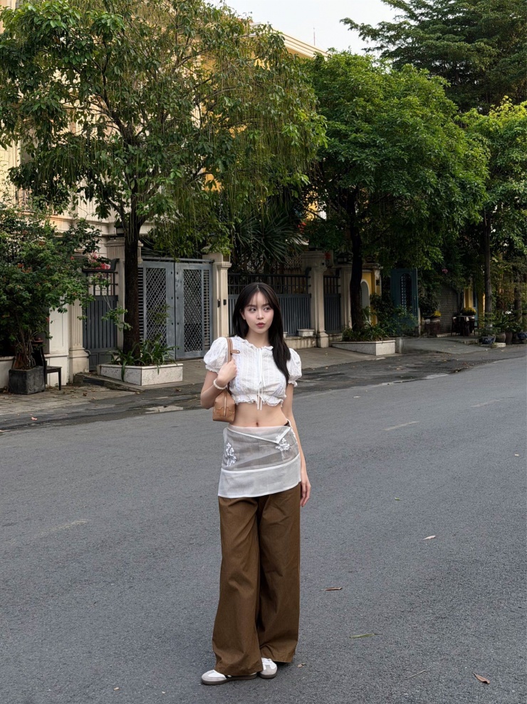 Chi tiết váy lưới mỏng layering bên ngoài càng làm bộ trang phục thêm phần mới mẻ, thể hiện gu thời trang có sự thử nghiệm nhưng không phô trương. Phụ kiện túi xách nhỏ gọn và giày sneaker đơn giản góp phần hoàn thiện hình ảnh năng động, gần gũi.