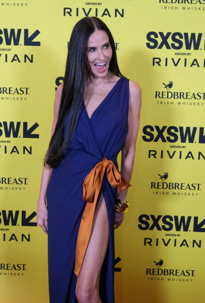 Nghệ sĩ 63 tuổi khoe dáng trong thiết kế của Saint Laurent - một chiếc váy quấn màu xanh với phần cổ khoét sâu và đường xẻ tà cao táo bạo. Ở phần hông, bộ trang phục được cố định bằng một chiếc nơ cam cỡ lớn.