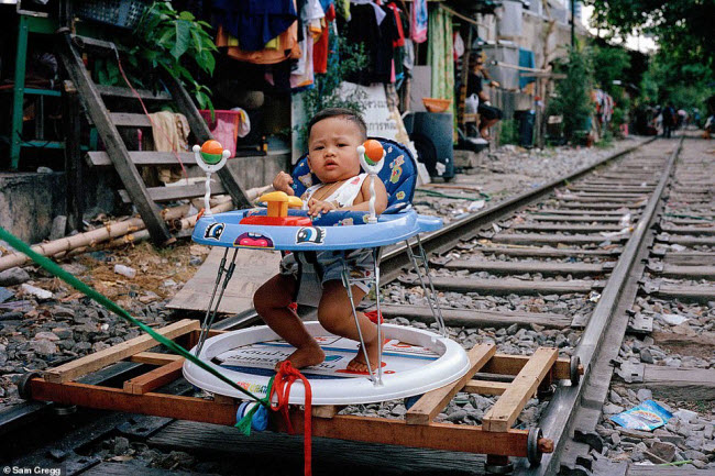 Em bé ngồi trong xe trượt&nbsp;được lắp bánh di chuyển trên đường ray tàu hỏa chạy qua khu ổ chuột.