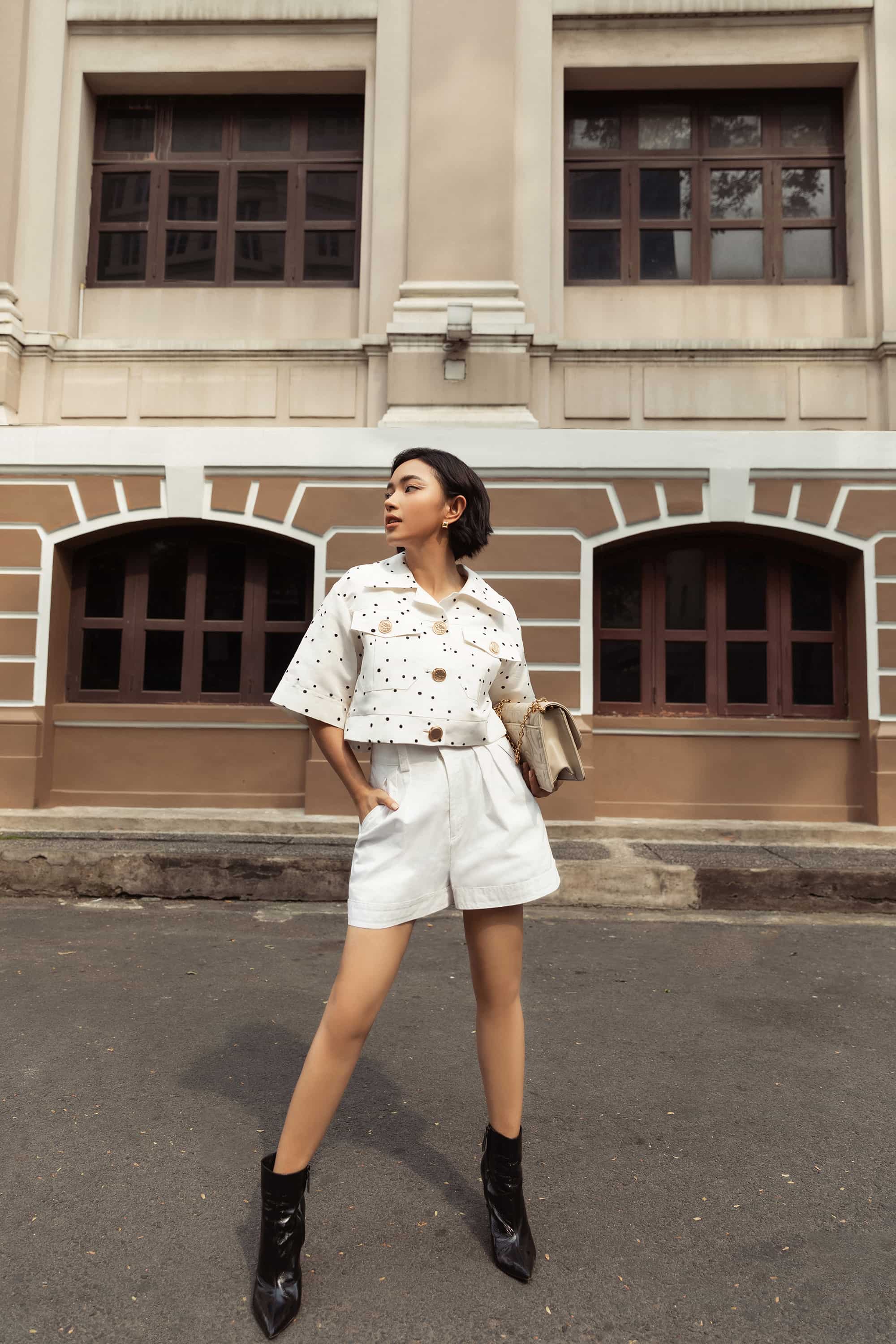 Cùng sử dụng quần short trắng nhưng Châu Bùi lại có cách phối hoàn toàn khác. Nàng fashionista phối cropped–jacket chấm bi cùng quần short để khéo ăn gian đôi chân. Châu Bùi phối cùng túi xách màu beige hợp tone cùng trang phục, mang đến cảm giác năng động trẻ trung. Điểm nhấn ở đôi boot da giúp cân bằng màu sắc cho tổng thể set đồ.