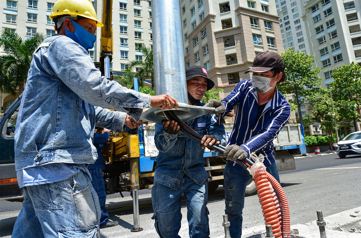 Công nhân điện lực tiếp tục lắp đặt những cột đèn chiếu sáng mới giữa dải phân cách tại đoạn đã hoàn thành mặt đường.