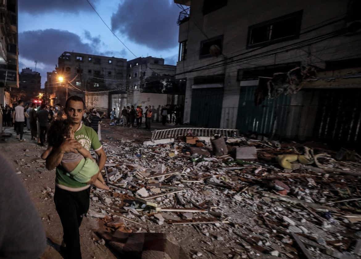 Người đàn ông ôm một đứa trẻ bước đi qua đống đổ nát ở Dải Gaza sau đợt không kích của Israel. Ảnh: Anadolu