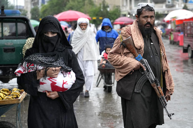 Phụ nữ Afghanistan đi bộ trên đường vào ngày 3.5.2022, bên cạnh là một tay súng Taliban.