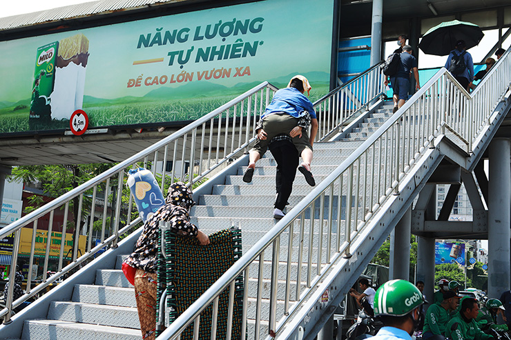 Người nhà bệnh nhân cõng bệnh nhân lên cầu vượt trước cổng Bệnh viện Bạch Mai trong thời tiết oi bức.
