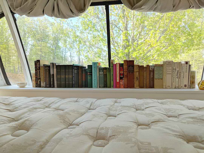 Phòng ngủ nhỏ nhắn ấm cúng với khung cửa sổ lớn và giá sách nhỏ.
