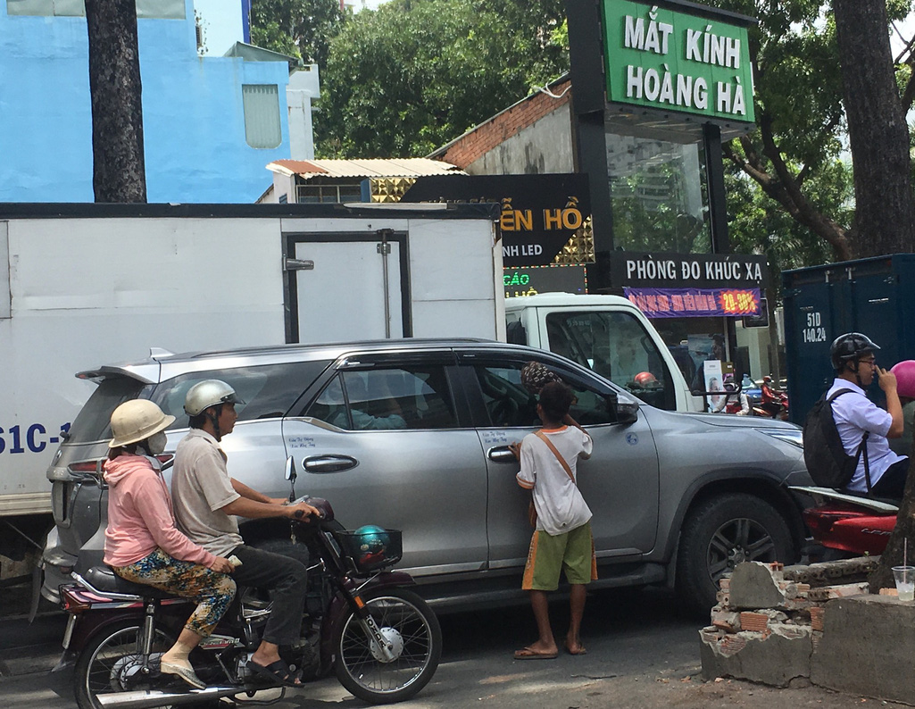 Đứa trẻ mặc áo cộc tay, đầu trần, giang nắng, kiên nhẫn xin tiền bố thí giữa dòng người đi xe máy và ôtô.