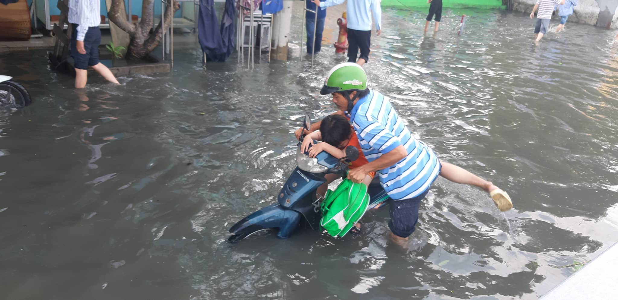 Phụ huynh “bơi” trong nước đưa con đến trường. Nước ngập khiến nhiều người phải bì bõm lội trong dòng nước đen ngòm, hôi thối ở khu vực đoạn bờ bao bị vỡ. (Ảnh: P.T.L.)