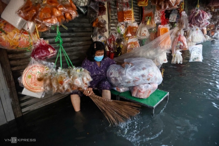 Một tiểu thương ở chợ Thủ Đức treo hàng hoá lên cao, vừa dùng chổi ngăn rác và nước tràn vào sạp hàng.