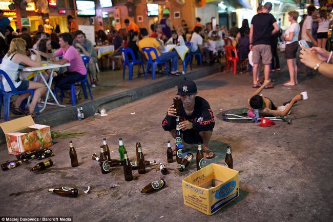 Người đàn ông biểu diễn với những chiếc vỏ chai bia trong khi một người tàn tật nằm ăn xin trên đường, cạnh rất đông du khách đang ngồi ăn uống.