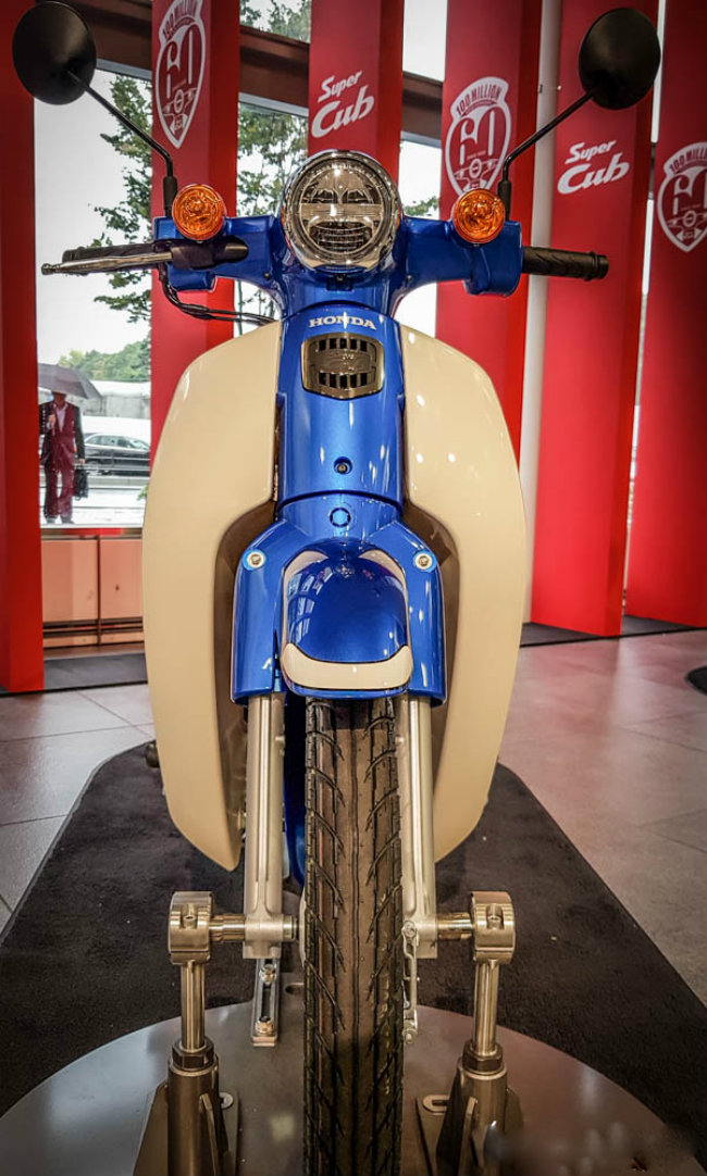 2018 Honda Super Cub 110 mang hệ thống phun nhiên liệu điện tử PGM-Fi và nút đề điện. Dung tích bình xăng đạt 4.3 lít và có cụm đồng hồ ly hợp ướt 4 cấp. Ảnh phía trước của 2018 Honda Super Cub 110 màu xanh sóng kim loại.