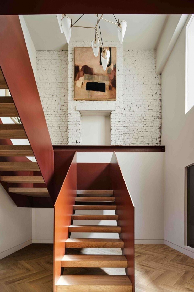 Cầu thang thép màu đỏ gỉ được thiết kế như một khối điêu khắc, kết hợp bậc gỗ sáng và sàn lát xương cá. Giải pháp này giúp cầu thang vừa là lối giao thông, vừa trở thành điểm nhấn thị giác xuyên suốt các tầng
