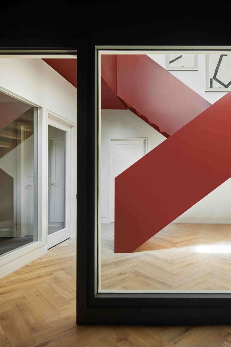 Góc nhìn qua khung cửa kính cho thấy cầu thang đỏ gỉ như một mảng màu trôi nổi trong không gian. Cách đặt cầu thang quanh lõi thông tầng giúp ánh sáng lan tỏa và tạo chiều sâu thị giác cho ngôi nhà